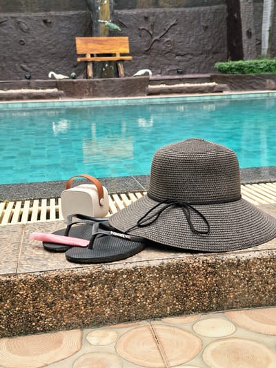 This is an outdoor, high-angle, full shot of swimming pool accessories laid out next to a swimming pool. The setting appears to be a tropical location, possibly Sihanoukville, Cambodia, given the context. The pool water is a vibrant turquoise, reflecting the bright light.
The primary subjects are a wide-brimmed straw hat, a pair of black flip-flops, a small, light-colored portable speaker with a brown strap, and a tube of lip gloss. The hat is dark brown with a black ribbon tied in a bow. The flip-flops are black and appear to be Havaianas. The speaker is positioned between the hat and the flip-flops. The lip gloss is pink and has text on it.
The objects are resting on a light-colored slatted surface, which is adjacent to a textured border of small, multicolored pebbles. Beyond the pebble border, there are large, circular tiles with a wood-grain pattern.
To the left of the pool, there is a dark, textured wall that resembles natural rock formations, with some greenery growing on it. A small, wooden bench-like structure is visible against this wall.
The scene suggests a moment of relaxation or preparation for swimming. The bright lighting indicates it is daytime, and the overall atmosphere is peaceful and leisurely.
Visible text includes "Havaianas" on the flip-flops and "Crystal Gloss" on the lip gloss tube.