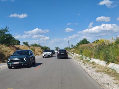 In Krushovitsa, Bulgaria, on a two-lane asphalt road, a caravan of vehicles travels under a bright blue sky dotted with white cumulus clouds. The leading vehicle, a dark green Kia Sportage, displays the license plate "TNB 125". Following closely behind are a white Toyota RAV4 with license plate "P 5378 MA", a black SUV, and a white hatchback. The roadside is flanked by dry, grassy vegetation and scattered bushes, with a power line tower visible in the distance. The scene captures a moment of transit on a rural Bulgarian road.