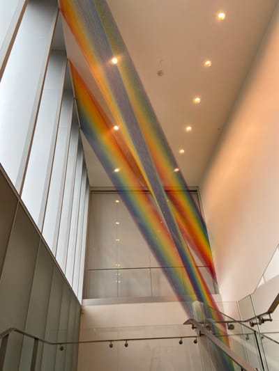 The image shows the interior of a modern building, likely a museum or art gallery. There are two large strips of rainbow-colored string art hanging from the ceiling, which is white and contains multiple recessed lighting fixtures. There is a glass railing and a staircase to the right, and a large window on the left, through which more string art is visible. The image is taken from a low angle, looking up at the ceiling. The location is likely in the United States, based on the building's architecture and the presence of string art, a popular art form in the country. The image is a representation of the string art installation, "Rainbow," by artist Olafur Eliasson, and the building is the The Shed in New York City.