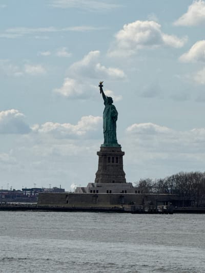The image shows the Statue of Liberty from a distance, positioned on the left side of the frame against a cloudy sky. The statue is green and stands atop a gray pedestal structure. To the right of the statue, the image captures a shoreline or a part of an island, with a row of trees and some buildings. The water body occupies the right portion of the image. The image seems to be taken on a clear day with the sun shining. While the specific location is not evident without additional information, it suggests a view across a body of water toward Liberty Island, which is visible from Jersey City, New Jersey.