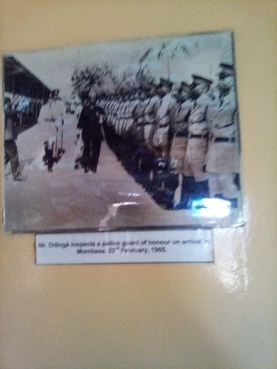 A framed, black and white historical photograph is mounted on a light beige wall. The photograph depicts an outdoor scene featuring a dark-suited individual, identified by the accompanying caption as Mr. Odinga, inspecting a police guard of honor. Mr. Odinga is positioned in the mid-ground, moving from left to right, appearing to review a long formation of uniformed police officers. These officers, dressed in light-colored attire and caps, stand in disciplined rows extending diagonally from the mid-ground towards the background on the right. Additional indistinct figures are present near Mr. Odinga and adjacent to a structure visible on the far left. Trees are discernible in the background. The caption directly below the image states: "Mr. Odinga inspects a police guard of honour on arrival in Mombasa, 23rd February, 1965." The overall image is slightly out of focus, and reflective glare is visible on the surface of the historical photograph, particularly in the lower right quadrant. The event occurred in Mombasa, Kenya, not South Sakwa ward.