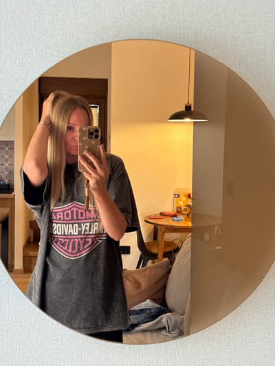 A woman is taking a mirror selfie in what appears to be a living room. She has long blonde hair and is wearing a dark grey t-shirt with a pink and white Harley-Davidson logo. The mirror is round and has a warm, bronze tint. In the reflection, a small wooden table with a red object and a box of cereal is visible. A pendant light hangs from the ceiling. The background shows a textured wall and hints of furniture. The location is identified as Kovačka Dolina, Montenegro.