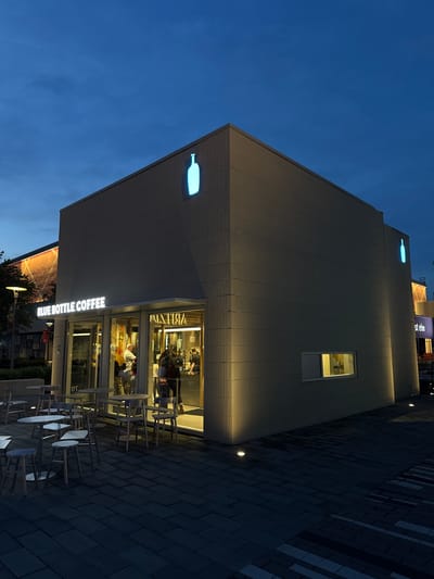 Captured from a tilted vantage point, the image presents the exterior of a Blue Bottle Coffee establishment. The building's facade is illuminated, highlighting the brand's logo. Tables and chairs are arranged outside the entrance, indicating an outdoor seating area. The setting appears to be an urban environment in Oak Brook, United States. Patrons can be observed inside the building. The sky transitions from light to dark, suggesting it is evening or dusk.