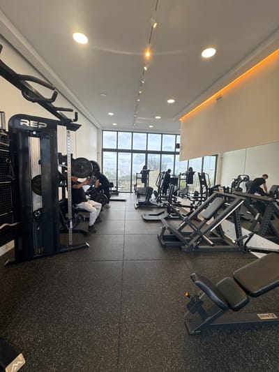 The image displays the interior of a fitness gym located in Plataniás, Greece. The facility features a dark grey rubberized floor with a speckled pattern, laid in square tiles. The walls are predominantly white, and the white ceiling incorporates multiple recessed circular lights and a linear track lighting system with spotlights. A section of the right wall includes a recessed light strip emitting an orange glow.
The far wall consists of large, floor-to-ceiling windows providing a view of an outdoor urban landscape, including buildings and trees under an overcast sky. The gym is equipped with various weight training machines. In the left mid-ground, a black "REP" branded multi-gym or power rack system is prominent. A male individual, dressed in a black top and white pants, is seated at this machine, engaging with loaded weight plates. Another person in a black shirt stands adjacent to him, appearing to assist or observe.
Further into the mid-ground and towards the right, multiple leg press machines and other weight machines are positioned. Several other individuals, mostly obscured or in silhouette against the bright windows, are visible utilizing various pieces of equipment. On the far right, one person is seen leaning over a machine. A black adjustable utility bench is partially visible in the right foreground.