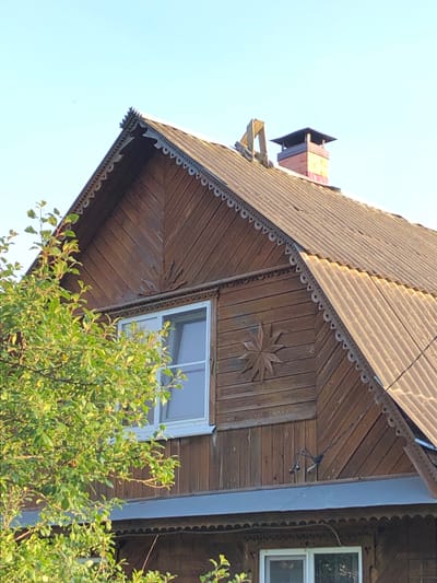 The image shows a close-up view of the upper part of a wooden house in Makeikha, Russia. The house features a gabled roof covered with corrugated material and adorned with decorative wooden trim along the eaves. There is a window on the upper level, and another window is partially visible on the lower level. A chimney made of brick with a dark cap rises from the roof. A tree with green leaves partially obscures the left side of the house. The sky is clear and blue.