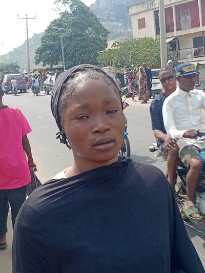 The image depicts a daytime street scene in Ozuri, Nigeria. A woman, positioned in the foreground, looks directly at the camera. She has dark skin, braided hair pulled back with a black headwrap, and wears a black draped garment over a white top, accessorized with black beaded earrings. To her left, another individual in a pink t-shirt and dark trousers walks away, carrying a dark bag. To her right, two men are seated on a motorcycle; the passenger wears dark sunglasses and a patterned shirt, while the driver wears a white collared shirt, shorts, green sandals, and a blue and yellow cap. The background shows a paved road with moderate vehicular and pedestrian traffic, including several motorcycles and cars. Various buildings, some multi-story, line the street on the right. Utility poles with overhead lines are visible. Dense green foliage and a large rocky mountain or hill are prominent in the distant background under a bright sky.