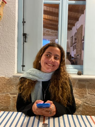 A young woman with long, curly brown hair, fair complexion, and a broad smile is seated at a table. She wears a black long-sleeved top, a light blue patterned scarf, silver hoop earrings, and a silver necklace with a dagger pendant. Several silver rings are visible on her fingers. She holds a blue-cased smartphone in both hands, positioned over a white tablecloth with blue and red stripes. A small metallic object, possibly a salt shaker lid, is present on the table in the foreground.

Behind the woman, a light blue-framed window is visible, with a black metal latch mechanism on its left side. The window reveals an interior space featuring a wooden beamed ceiling, a white wall, and a section of wall constructed from reddish-brown bricks or stones. Framed items are mounted on the white wall. Below the window is a rustic stone wall section, and a small, dark, spherical object rests on the window sill. To the far left, a portion of a decorated ceramic pitcher with orange and red accents is visible on a textured light-colored wall. The scene depicts an indoor dining environment, characteristic of a traditional restaurant or taverna, located in Megalochori, Greece.