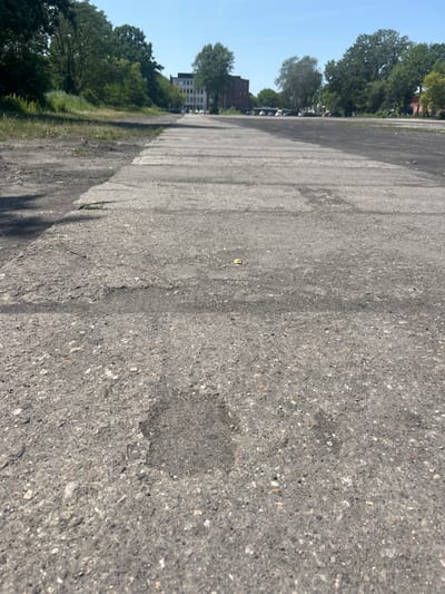 This is a low-angle, close-up photograph of a cracked and weathered concrete surface, likely a road or parking lot, with a faint shadow cast on the left side. The concrete is uneven, with patches of darker, rougher texture. In the foreground, a small, circular depression is visible.
In the midground, the concrete surface extends into the distance, showing parallel lines of concrete slabs that form a path or road. To the left of the concrete, there is an area of dirt and sparse, dry grass. Further back, a line of trees borders the area, with some buildings visible behind them. One building appears to be a multi-story structure with a red brick facade, while another is lighter in color and has many windows. A few cars are parked along the road in the background. The sky above is clear and blue.
The scene appears to be an outdoor urban or semi-urban environment, possibly a disused industrial area or a less-maintained public space. The overall impression is one of an aging and somewhat neglected infrastructure. The specific location in the city of Konstancin-Jeziorna, Poland, is not evident from the visual information within the image itself, but this description is provided as per the prompt's instruction. There are no people visible in the image.
