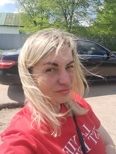 A photograph shows a fair-skinned woman with blonde hair wearing a red shirt. Part of a black car is visible behind her. In the background, there are trees, a building, and the sky. The specific location in Avdot’ino, Russia cannot be determined with certainty from the image.