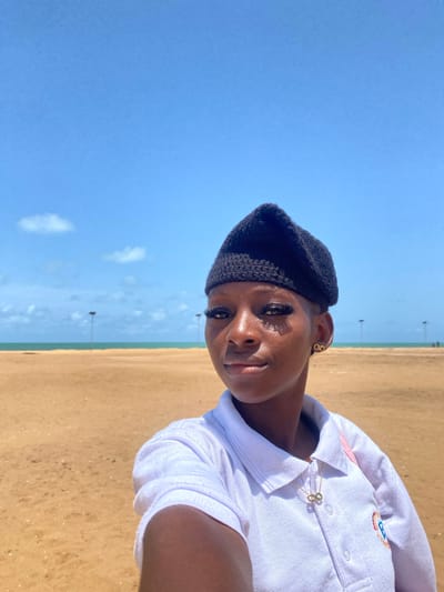 A young woman stands on a beach in Djondji, Benin, under a clear blue sky with a few scattered clouds. She is wearing a black beret and a white collared shirt with an infinity symbol pendant. The beach consists of tan sand that stretches to the sea. In the background, evenly spaced poles are visible near the water's edge.