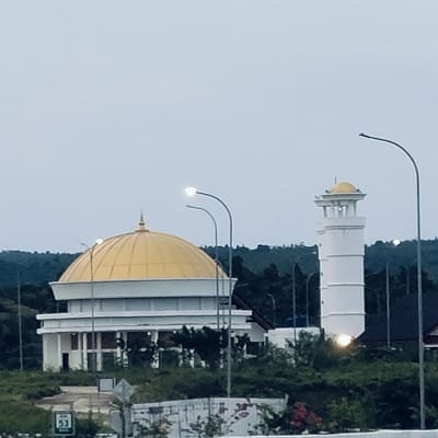 The image displays a serene architectural scene of a mosque complex in Cotkareung, Indonesia, under an overcast sky. The central feature is a white mosque building, topped with a large, gleaming golden dome. Its facade reveals multiple levels and structural pillars or columns.
To the right of the mosque, a tall, slender white minaret stands prominently. This minaret also features a smaller golden dome at its apex, with an open, columned section below it. Several streetlights are visible, with their lamps brightly illuminated, suggesting either dawn, dusk, or a heavily overcast day. One streetlight arches gracefully between the mosque and the minaret, while another appears closer to the minaret.
The buildings are surrounded by abundant green foliage and trees, forming a dense, natural backdrop that contrasts with the white and gold structures. The setting appears peaceful and without any direct human activity. In the bottom left corner of the image, a small green and white sign is partially visible, showing the text "ATM" and the number "53."