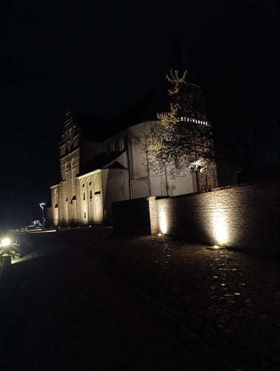 The image depicts the St. Aldegundis Church in Emmerich on the Rhine, Germany, at night. The church, constructed of brick, is illuminated by spotlights that highlight its architectural features, including its tower and windows. A brick wall runs along the right side of the frame, also illuminated by spotlights. A cobblestone path or road leads towards the church. At the start of the road on the left are two headlights. The overall environment is dark, with the only light sources coming from the spotlights and the headlights. A bare tree stands to the right side of the building.