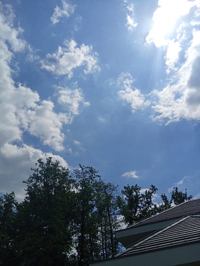 The image depicts a clear blue sky with scattered white clouds, characteristic of a sunny day. The sun is partially obscured by clouds in the upper right portion of the frame, casting bright rays. A small airplane is visible in the distance, flying through the sky. In the lower part of the image, the dark green canopy of trees creates a natural silhouette against the sky. A modern roofline, likely belonging to a building, with dark gray tiles and sharp angles, is visible in the foreground, spanning the lower right quadrant. The scene appears to be an outdoor setting, possibly in a suburban or rural area, given the presence of trees and a residential-style roof. The location is identified as Nowa Wieś Szlachecka, Poland. There are no people or interactions visible in the image.