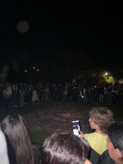 At night in Zidarovo, Bulgaria, a gathering of approximately 50 people stands in a line formation. Most face forward with limited interactions. The group is illuminated by overhead lighting and the glow emanating from what appears to be a ground-level fire or ember bed. In the foreground, individuals are focused on a cell phone screen, observed closely by others. The setting is outdoors, likely on grass or a similar natural surface.