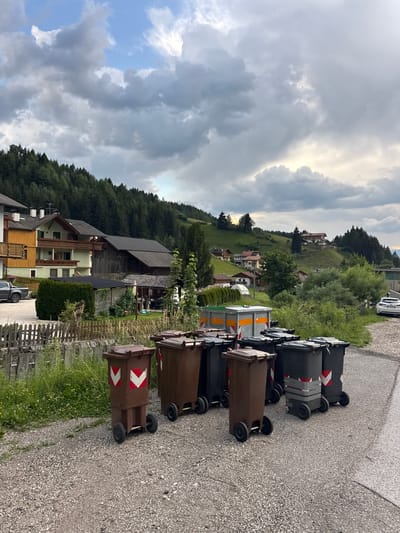 The image captures a view of St. Magdalena - Santa Maddalena, Italy, under a cloudy sky. In the foreground, a row of assorted trash bins is visible, including brown and black wheeled containers, some with red and white markings. Behind the bins, a gravel pathway leads towards a village nestled on a hillside, characterized by traditional alpine architecture with wooden balconies and dark tiled roofs. The houses are scattered amongst lush green trees and rolling hills, with a dense forest covering the upper slopes of the mountains. The overall scene suggests a quiet, rural setting in the Italian Alps.