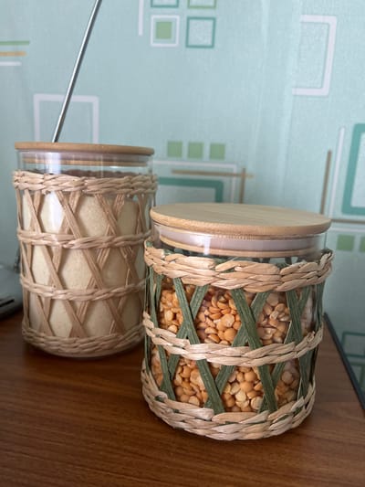 This media file features two glass jars with woven casings and bamboo lids, placed on a wooden surface. The top jar is filled with yellow split peas, and its woven casing is a mix of natural straw and green reeds. The bottom jar contains a light-colored granular substance, possibly sugar or flour, and its woven casing is entirely made of natural straw. Both jars have clear glass bodies and are topped with natural bamboo lids. The background shows a wall with a light blue geometric pattern. The lighting suggests an indoor setting, possibly a kitchen or pantry, with no indication of the time of day or weather. There is no visible text in the image. The overall scene is static, focusing on the decorative storage jars.