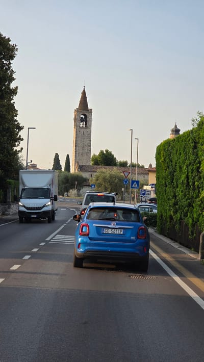 The image depicts a street scene in Bardolino, Italy, with traffic in motion. In the foreground, a blue Fiat 500X is seen from the rear, driving in the right lane of a two-lane road. The car's license plate reads "CD 541LP". To the left of the Fiat, a white Iveco delivery truck is visible in the adjacent lane. The road has white dashed lines separating the lanes and a white pedestrian crossing.
In the background, a tall, stone bell tower with a conical roof rises above buildings and trees. To the right of the bell tower, a smaller domed structure is partially visible, also suggesting religious architecture common in Italian towns. A dense, green hedge lines the right side of the road. Traffic signs, including a blue "P" indicating parking and a circular sign with a red border and diagonal line suggesting a prohibition, are present on the right. The sky is clear and a soft, golden light suggests it is either early morning or late afternoon. The overall environment appears to be a well-maintained urban or semi-urban area.