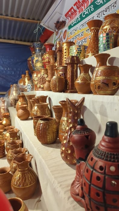 A display of earthenware pots and vases, arranged in rows. The ceramics are primarily brown, with some displaying intricate carvings, gold accents, or decorative mirrors. A red and black wind instrument is also visible. A banner with text is partially seen in the background.