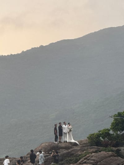 The image captures a wedding photoshoot underway on rocky terrain in Sơn Trà Ward, Vietnam. In the upper mid-ground, a couple, presumably the bride and groom, stands prominently on an elevated rock formation. The bride is dressed in a white wedding gown, trailing to her right, and holds a white floral bouquet. The groom stands beside her in a white suit. To their left, two men in dark, casual attire (black shirts, shorts, and caps) stand near the couple, appearing to be crew members.

In the lower foreground, multiple individuals are positioned on a lower cluster of rocks. At least two individuals are visible with cameras; one, wearing a black shirt with the number "10" on the back, is crouched and operating a camera, directed upwards towards the bridal party. Another person in a light-colored top stands nearby. On the far right, a small cluster of green foliage is visible, with what appears to be part of a light stand or equipment rig partially obscured behind it.

The background is dominated by a vast, steeply sloped, and densely forested mountain, exhibiting a muted green-grey tone. The sky above is light and hazy, indicating an overcast day or specific light conditions. The overall environment consists of large, rugged, grey-brown rock formations.