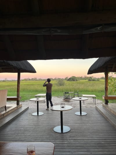 A man stands on a wooden deck in Khwai, Botswana, overlooking a grassy field under a pastel sky, possibly at sunset. The scene is partially obscured by a thatched-roof building on the left, with several round tables and chairs positioned on the deck, offering a view of the landscape. The man, wearing a brown shirt and dark pants, is facing away from the viewer, seemingly observing or photographing the scenery.