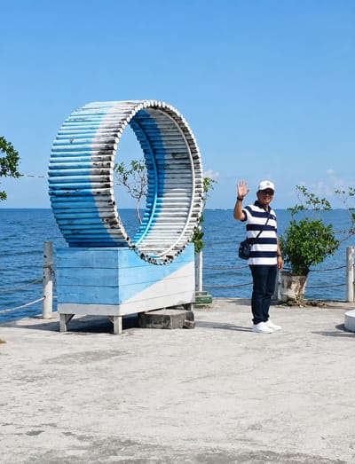 A person is standing on a concrete pier by the ocean at Pantai Cermin Kiri, Indonesia. The person is wearing a striped polo shirt, dark pants, white sneakers, and a white cap. They are waving their right hand and have a dark bag slung over their shoulder. To the left of the person is a large, circular, blue and white wooden structure supported by a blue and white wooden base. The ocean is visible in the background, with a clear blue sky above. There are also some green plants and a white railing on the pier.