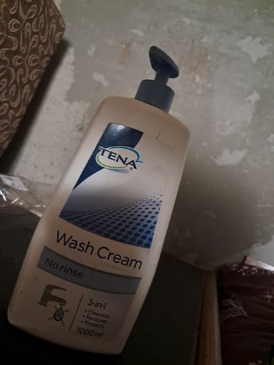 A TENA Wash Cream bottle, 1000 ml, is placed on a dark surface. The bottle is mostly off-white with a blue and white label. The label displays the TENA logo, "Wash Cream," "No rinse," and "3-in-1: Cleanses, Restores, Protects." A graphic of a faucet with a water droplet crossed out is visible on the lower left of the label. The background consists of a textured, light-colored wall with some discoloration and a patterned fabric on the left side. The scene is indoors, likely a bathroom or storage area. The specific location of Chobruchi, Moldova is not discernible from the visual information provided in the image.