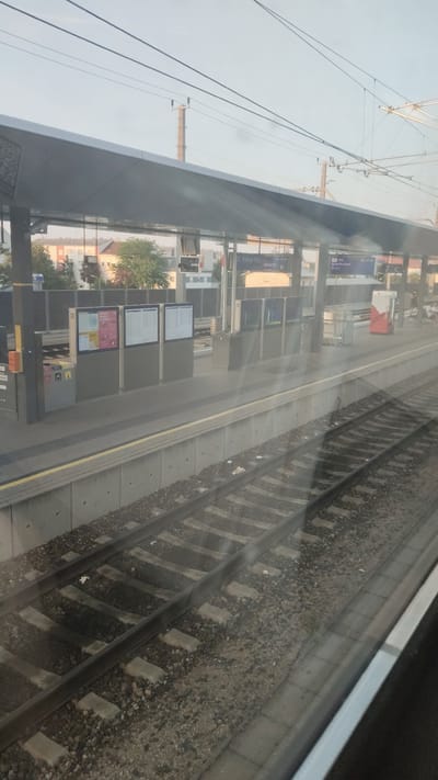 This media file captures a view from a train window looking out at a train station platform. The scene is set at Sankt Pölten, Austria. The time of day appears to be late afternoon or early evening, as indicated by the soft, angled sunlight casting long shadows and creating a warm glow on the buildings and platform. The sky is clear, suggesting fair weather.

The main subjects are the train station platform, railway tracks, and overhead power lines. The platform is covered by a modern, grey, cantilevered roof supported by columns. Beneath the roof, there are several digital display boards showing train schedules. One prominent display clearly reads "St. Pölten Hbf" and lists departure times, including "20:10:34" and "20:10:35." Another display shows information for a train labeled "RJ 712" departing at "21:39 Pöchlarn." Several trash receptacles are visible on the platform, along with what appear to be information kiosks or ticket machines. In the background, residential buildings and trees are visible beyond the station. A red and white vending machine or kiosk stands on the far right of the platform.

The railway tracks, with their gravel ballast and concrete sleepers, run diagonally across the foreground and mid-ground of the image. The train is stationary, implying that this is a stop at the station, and the activity is the normal operation of a train station. There are no people clearly visible in the frame, but their presence is implied by the infrastructure of the station. The overall mood is calm and functional, typical of a transit hub. The sunlight reflecting on the window creates a slightly hazy or ethereal quality to the image.