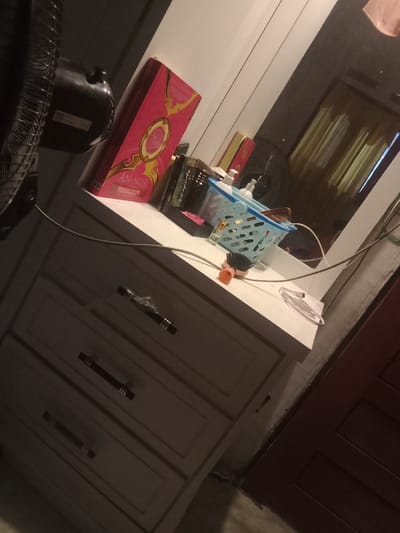 A dresser with three drawers sits near a red door. An assortment of items rests on top of the dresser, including a pink box labeled "AN-NUR," containers, a blue basket, and an orange-handled brush. A portion of a fan is visible in the image's left. A mirror behind the dresser reflects a room with curtains. A white power cord is also draped across the scene.