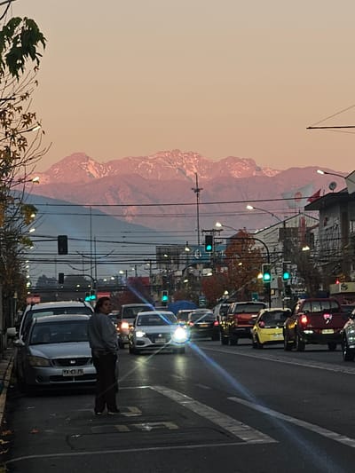 The image depicts a street scene in San Fernando, Chile, during a twilight hour, with snow-capped mountains visible in the distance. The sky is a soft gradient of pink and pale orange. In the foreground, a person stands on a pedestrian crossing, looking to the left. They are wearing dark pants and a grey hooded sweatshirt. Several cars are on the street, including a silver sedan on the left with license plate "TX-85-75". A line of vehicles is stopped or moving slowly on the right side of the road. Traffic lights, some displaying green, are visible throughout the scene. Buildings line the right side of the street, some with advertisements. Overhead power lines and streetlights are prevalent. Trees with sparse autumn foliage are present on the left side of the image.