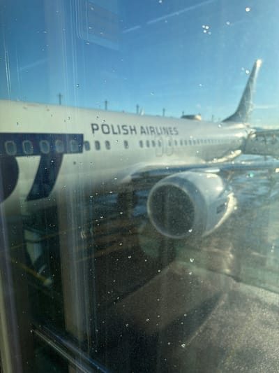 An airplane with "POLISH AIRLINES" written on its side is visible through a window with water droplets on it. The airplane is white with a dark blue logo on the side. It is parked at an airport gate, with a jet bridge attached. The sky is blue and clear, and the sun is shining. This image was taken at Oslo Airport, Gardermoen, Norway.