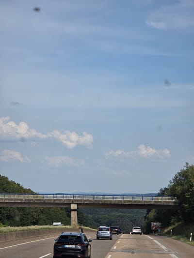This is a vertical, eye-level, medium shot taken from a vehicle traveling on a highway in Windhagen, Germany. The sky is blue with scattered white clouds. In the foreground, a black Skoda Octavia is traveling in the right lane, displaying the license plate "EU VA 2981". To the right of the Skoda, a portion of a gray vehicle is visible in the same lane. In the left lane, a blue Skoda Fabia is visible, followed by a dark colored SUV. Further ahead, a white SUV is in the right lane. A highway overpass stretches across the scene, supported by concrete pillars. The overpass has a yellow railing on the bottom and a blue and white railing on top. Dense green trees and vegetation line the sides of the highway and surround the overpass. A signpost with an image and the text "Wiedtal 44m" is visible on the right side of the road.