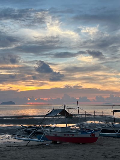 This is a vertical, eye-level, medium shot of several outrigger boats pulled up onto a sandy beach at dawn in Cuyo, Philippines. The sky is a dramatic display of clouds painted with hues of orange, pink, yellow, and blue, indicating the rising sun. The sun itself is a bright orange glow just above the horizon, peeking through a break in the clouds. The ocean is calm and reflects the colors of the sky. In the distance, a small island or landmass is visible on the left. The boats are traditional Filipino outriggers with bamboo outriggers and canvas coverings. One boat in the foreground is painted red and white with "ROLYN" and the number "2025" in red lettering. Another boat behind it is partially visible and appears to be dark blue. The beach is composed of sand and what looks like mudflats exposed by the low tide. There are no people visible in the image. The overall atmosphere is serene and peaceful, capturing the beauty of a tropical sunrise.