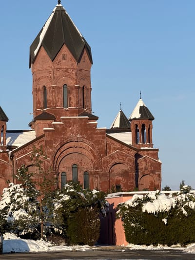 This image displays a large, reddish-brown stone church located in Rassvet, Russia, under a clear blue sky during winter. The primary structure is dominated by a central octagonal tower with a dark, pointed, pyramidal roof exhibiting significant snow accumulation on its upper facets. This tower features multiple tall, narrow arched windows. The main body of the church below the central tower is constructed from the same reddish stone, characterized by large, decorative rounded arches above sections containing smaller arched windows. To the right, two smaller auxiliary towers, also topped with dark, conical roofs partially covered in snow, are visible. The ground in the foreground is uniformly covered in white snow. Numerous evergreen trees and bushes, heavily laden with snow, occupy the lower left and mid-ground. A section of a light pink wall or building is partially visible behind the snow-covered vegetation on the right side of the frame. The scene is brightly illuminated by natural light. No individuals or active subjects are present.