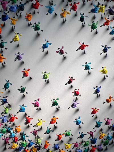 The image displays a close-up, overhead view of a white, textured surface adorned with numerous small, colorful, figurative sculptures. These sculptures, appearing to be made of thick paint or a similar pliable material, represent simplified human figures in dynamic poses, suggesting movement or dance. Each figure has a black, circular head. They are arranged in a scattered, seemingly random pattern across the surface, with some figures clustered together and others more isolated. The lighting casts distinct shadows from each sculpture onto the white background, indicating the direction and intensity of the light source. The overall impression is one of playful, vibrant energy conveyed through the multitude of brightly colored figures. There is no identifiable location within the city of Honfleur, France, visible in this image.