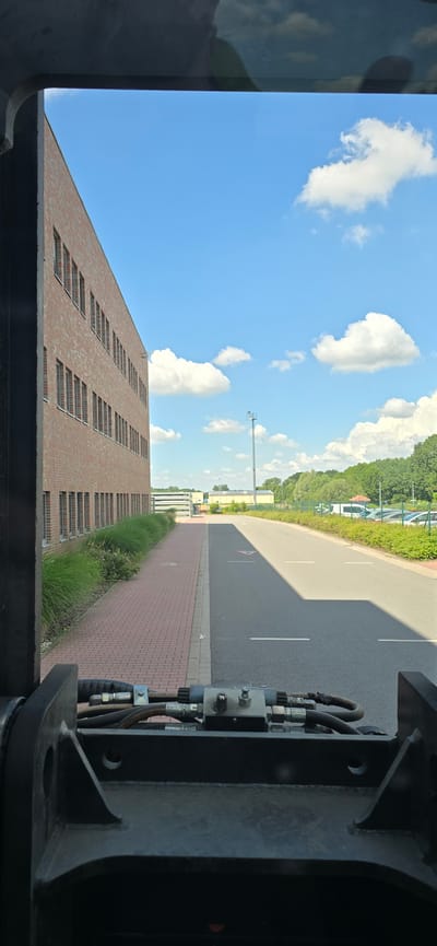 The image presents a view from inside a vehicle looking out towards a modern brick building on the left and a paved road on the right. The building has numerous windows and is surrounded by tall green grass. The road is bordered by a red brick sidewalk and further right by a grassy verge with trees in the distance. The sky is blue with scattered white clouds. In the foreground, the mechanical components of the vehicle, likely a loader or similar heavy machinery, are visible, including hydraulic hoses and connectors. The scene appears to be a commercial or industrial area on a bright, sunny day. This location is identified as being in Haren (Ems), Germany. There are no people clearly visible in the image.