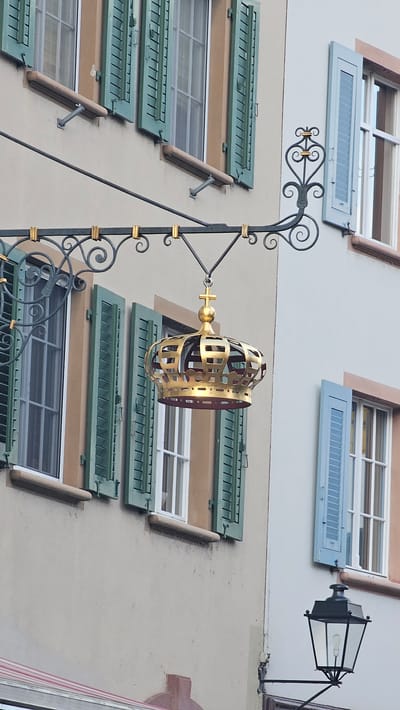 A medium shot shows a beige building with two windows on the upper story and three on the lower story. All the windows have exterior shutters with louvers. The upper windows have pale-blue shutters that are open, while the lower windows have dark-green shutters that are open.

A sign consisting of a gold, cage-like crown suspended from a black-iron bracket is visible on the wall between the upper and lower windows. The sign is suspended from a wrought-iron pole that is attached to the building. A vintage lamppost is visible near the upper-right corner of the frame. The location is likely Rheinfelden, Switzerland, based on the style of the architecture and the crown in the sign.