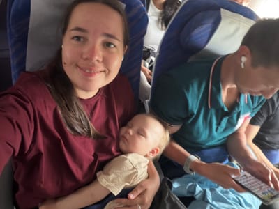The image shows a group of people on an airplane. In the foreground, a woman is holding a sleeping baby. The woman is smiling and looking at the camera. The baby is wearing a light-colored onesie. To the right of the woman, a man is looking at his phone. He is wearing a teal polo shirt and white earbuds. The background is out of focus but shows airplane seats and windows. The scene takes place inside an airplane, possibly during a flight to or from Ota, Japan.