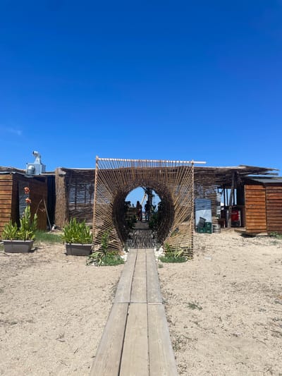 The image depicts an outdoor establishment, likely a beach bar or restaurant, situated in Ierissos, Greece. A wooden pathway leads towards an arched entrance constructed from bamboo. Beyond the entrance, several individuals are visible, appearing to be patrons. The establishment itself features rustic structures made from wood and bamboo, with various objects like crates, kegs, and a refrigerator visible. Green plants in rectangular planters are strategically placed around the entrance, adding a touch of greenery to the sandy environment. The sky above is a clear, vibrant blue.