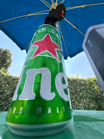 A close-up, low-angle shot shows a condensation-covered can of Heineken beer positioned on a green, reflective surface, possibly a table. The can is tilted, with the large white lettering of the brand name prominently displayed. Behind the can, a large blue umbrella provides shade, and a black baseball cap with a red logo is attached to its central pole. To the right of the can, a dark gray, rectangular object with visible text and a light colored border is present, suggesting it might be an electronic device or speaker. The background reveals a lush green hedge against a clear blue sky with some sunlight filtering through. The overall setting appears to be outdoors, likely in a garden or patio area, with no people visible in the frame.