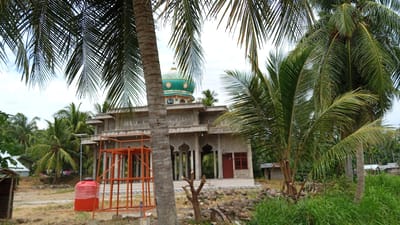 This is a frontal, medium-range, eye-level shot of a concrete building with a green dome, resembling a mosque, surrounded by lush vegetation and palm trees in Cotaneukbatee, Indonesia. The building has a light gray facade with arched doorways and windows, and intricate decorative patterns along the upper sections. A bright red water tank is situated to the left of the building, next to an orange metal structure that appears to be scaffolding or a stand. The ground is uneven and rocky, with patches of dry earth and grass. In the background, more palm trees and smaller buildings are visible. The sky is overcast, suggesting a cloudy day. The scene appears to be a construction site or a newly built religious structure in a tropical, possibly rural, environment. No people are visible in the image.