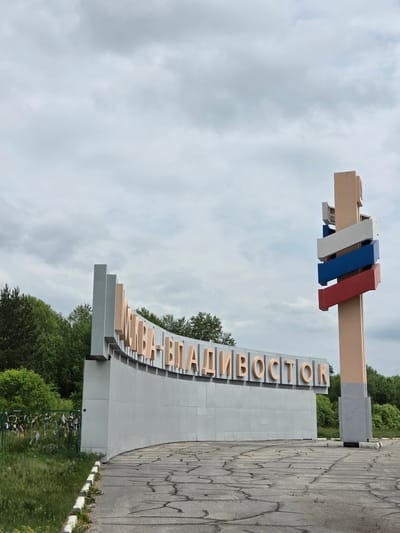 The image shows the entrance sign to the city of Nikolaevka, Russia. The sign is large and curved, with the name "Николаевка-Владивосток" (Nikolaevka-Vladivostok) spelled out in large, light brown letters. To the right of the main sign is a tall, slender tower with horizontal rectangular panels in the colors of the Russian flag: white, blue, and red. The ground in front of the sign is paved with cracked asphalt, and there is a grassy area to the left with a green metal fence adorned with numerous colorful ribbons or fabric strips tied to it. Trees and foliage are visible in the background, and the sky is overcast.