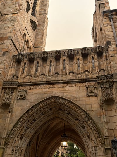 This image shows an exterior view of a gothic-style building, likely part of a university, with intricate stone carvings and architectural details. The building is constructed with rough-hewn, light brown stones, some of which appear to be sandstone.
In the foreground, a large, arched entryway is visible. The arch is elaborately decorated with Gothic tracery, including carved foliage and small figures. Above the arch, a stone frieze runs horizontally, bearing what appears to be Latin text carved in a Gothic script. Below the frieze, on either side of the entryway, are carved escutcheons or coats of arms. One escutcheon depicts a book open at the top, with a lamp or oil vessel below it and a sprig of leaves.
Above the frieze, a decorative stone balcony or walkway extends across the building. This structure features multiple small, arched windows and ornate corbels that support the walkway. The stonework here is detailed and layered.
The building rises upwards on either side of the frame. On the left, a portion of a tall tower with pointed windows is visible. On the right, another section of the building shows a similar stone construction and architectural style.
Suspended within the arch of the entryway is an ornate, black metal lantern with glass panes, illuminated from within. Through the arch and beyond the lantern, greenery and trees are visible, suggesting an outdoor setting. The sky above is overcast and pale.
The architectural style and inscription suggest this building is likely a historical academic institution. Based on the inscription "FOR GOD AND COUNTRY" visible on the frieze and the gothic architecture, this building is the Harkness Tower at Yale University in New Haven, Connecticut.