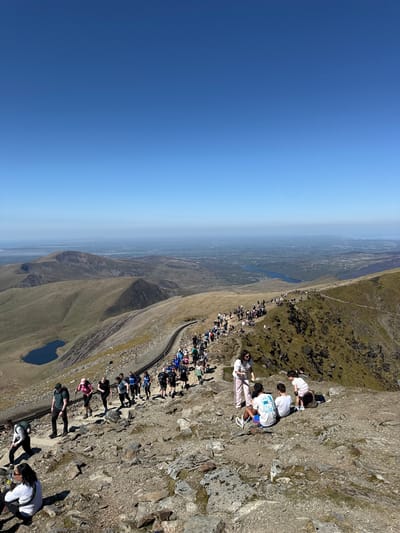 The image shows a landscape, likely in Beddgelert, United Kingdom, with a steep, rocky terrain on the right side and a distant view on the left under a clear blue sky. A group of people are situated on the rocky terrain, some standing, some sitting. A railway track runs along the edge of the rocky area. In the distance, there is a lake or body of water.