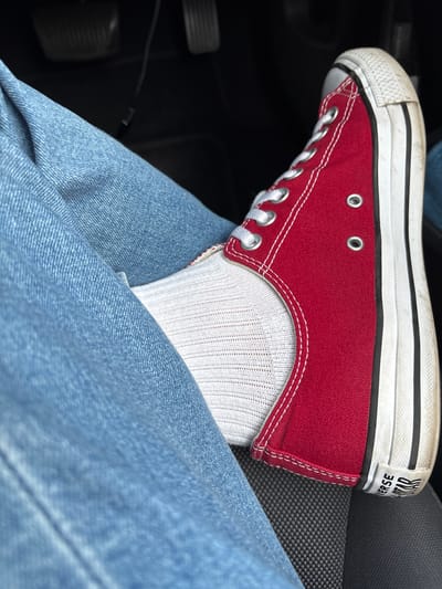 The image shows a close-up of a person's foot wearing a red Converse sneaker and white socks, with blue denim jeans visible. The sneaker has white laces and a white sole with black stripes. The person appears to be sitting, possibly in a car, as indicated by the presence of a dark, textured surface which could be a car seat, and a blurred dark background that might be the car's interior. The lighting suggests an indoor or shaded outdoor environment. There is no visible indication of the specific location within the city of South Staffordshire, United Kingdom.