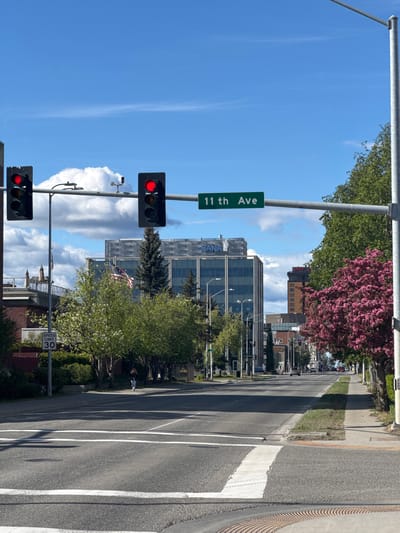 The image shows a street scene in Anchorage, Alaska, USA. The foreground is dominated by a paved road with white crosswalk markings. A speed limit sign indicating "30" is visible on the left side of the road. In the middle ground, there are traffic lights showing red, a street sign indicating "11th Ave", trees with green and pink blossoms, and several buildings, including a modern glass-fronted building with "NANA" visible on its facade. An American flag is also visible on a flagpole. The background shows more buildings and trees, with a blue sky and scattered white clouds above. A person is walking on the sidewalk on the right side of the street.