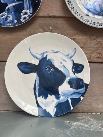 A white ceramic plate with a blue and white painting of a cow's head is displayed against a wooden background. The cow's head is painted in a watercolor style, with varying shades of blue and white. The cow has large, expressive eyes, prominent horns, and a black and white patterned coat. The plate is positioned slightly to the right of the center of the frame. In the background, two other plates are partially visible, one with a floral blue and white pattern and another with a blue and white patterned border. The wooden background appears to be made of planks. The scene is lit from above, casting a slight shadow on the plate. The overall impression is of a decorative display of ceramic art, possibly in a shop or home setting. There is no information to suggest the location is in Tuk, Netherlands.