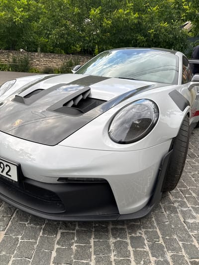 The image shows the front quarter of a silver Porsche 911 GT3 RS parked on a cobblestone street in the city of Morovaia, Moldova. The car has black racing stripes on its hood and fender flares. A license plate is visible on the front bumper. The background shows a stone wall and green trees. The lighting suggests it is daytime. No people are clearly visible in the scene.