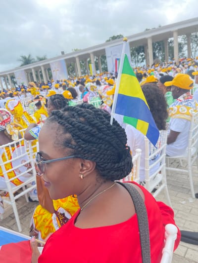 The image shows a gathering of people in what appears to be a public event, possibly related to a religious gathering, located in Mongomo, Equatorial Guinea. A woman with braided hair and wearing glasses is seated, partially facing away from the camera. She is wearing a red top and has a strap across her shoulder. A small Gabonese flag is positioned behind her head. The background features a large crowd of people wearing yellow and white clothing, many with matching hats. They are seated in white plastic chairs. In the far background, there is a building with architectural columns, possibly a civic or religious structure. The sky above the building is overcast. Banners with a cross can be seen behind the crowd. The setting suggests a significant public gathering.