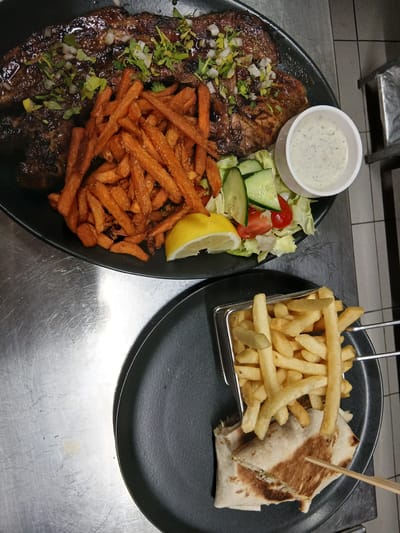 The image shows two black plates of food on a metallic surface. The top plate contains a large piece of grilled meat, possibly steak or pork, topped with finely chopped white onion and green herbs. It is accompanied by a pile of sweet potato fries, a small side salad with lettuce, cucumber, and tomato, a lemon wedge, and a small white bowl of dipping sauce, likely tzatziki or a similar yogurt-based sauce. The bottom plate features a portion of french fries in a small metal basket, served alongside a halved wrap or burrito that appears to be filled with meat and vegetables. The setting appears to be a restaurant kitchen or food preparation area, given the metallic surfaces and what seems to be stainless steel equipment in the background. The lighting suggests an indoor environment. The image does not provide any specific visual cues to pinpoint its location in Sotira, Cyprus.