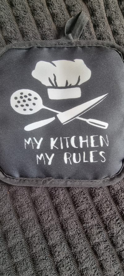 A close-up, high-angle shot features a black pot holder with a white graphic and text. The pot holder is resting on a dark grey, ribbed towel. The graphic on the pot holder depicts a chef's hat, a slotted spoon, and a knife, arranged as if for cooking. Below the graphic, white text reads "MY KITCHEN MY RULES". The pot holder has a small loop at the top. The background is a textured, dark grey towel. The image is well-lit and in focus, showcasing the details of the pot holder's design and the texture of the towel. There are no people or actions visible in the image. The scene is a still life, focusing on kitchen accessories. The image is not located in Naaldwijk, Netherlands.